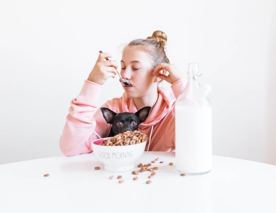 girl eating bowl of cereal with dog next to glass milk jug