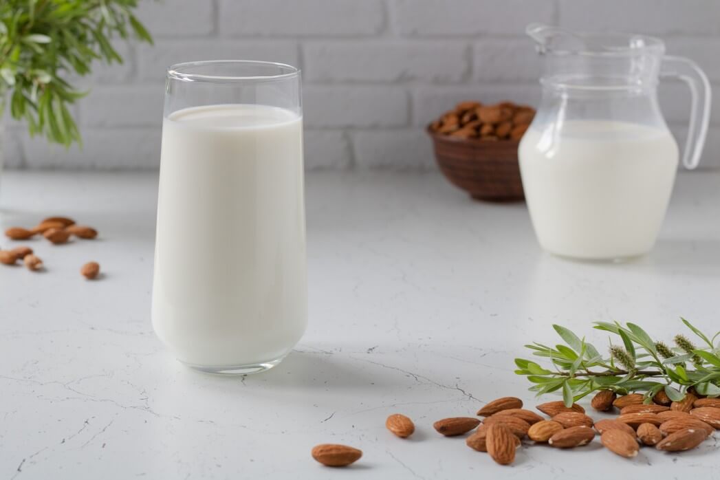 A glass of almond milk next to it is a bowl of almonds a jug almonds and sprigs of greens