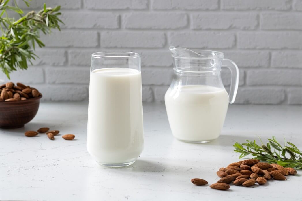Glass of almond milk next to a carafe, almonds and green twigs