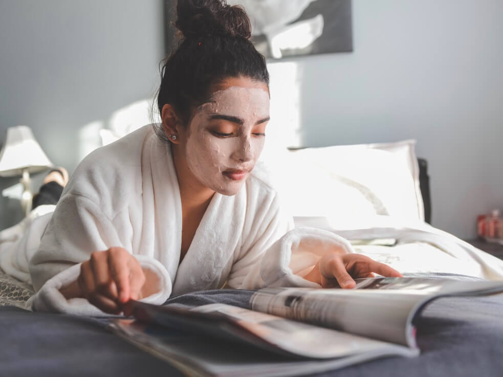 woman with facial mask reading magazine in robe on bed