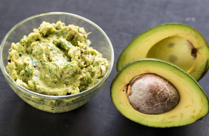 guacamole in clear bowl next to avocado with seed