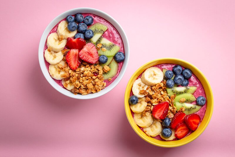 two acai bowls topped with fruit on pink background